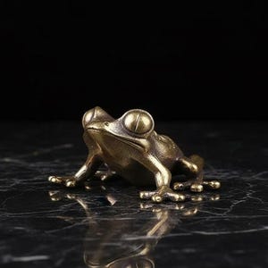 May include: A small, brass frog ornament with prominent, round eyes and outstretched legs. The frog is placed on a dark, reflective surface, producing a mirrored reflection. The metallic finish of the frog gives it a vintage or antique look.
