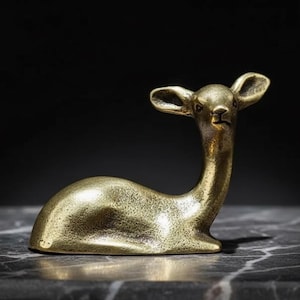 May include: A small, golden-colored deer figurine in a resting pose. The sculpture has a smooth, polished finish and is set against a dark background. The deer's antlers are small and its body is streamlined.