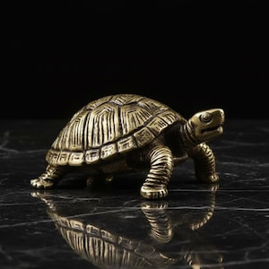 Peut inclure: Petite figurine de tortue en laiton doré, riche en détails. La carapace est texturée et la tortue est en position de marche. Le fond sombre met en valeur sa couleur dorée et son design complexe.