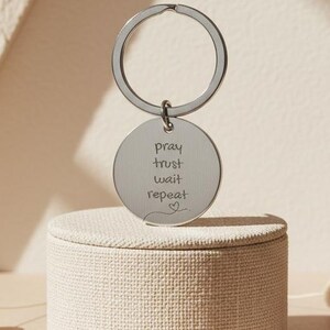 Può includere: Un portachiavi color argento con un ciondolo rotondo. Il ciondolo è inciso con le parole "pray, trust, wait, repeat" e un simbolo a forma di cuore. Il portachiavi è esposto su una piccola scatola rotonda beige.