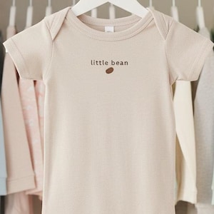 May include: A beige baby onesie with the text "little bean" and a brown bean graphic is hanging on a white hanger. The onesie is surrounded by other baby clothes in pastel colors, including pink, blue, and cream, hanging in a closet.