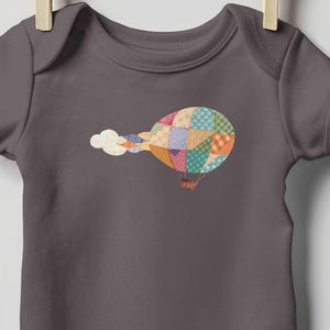 May include: A gray baby onesie with a colorful hot air balloon design. The balloon is made of patchwork squares in various colors and patterns. A cloud of white smoke trails behind the balloon.