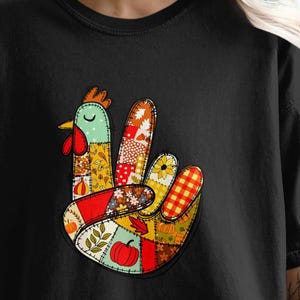 Puede incluir: Camiseta negra con un diseño en forma de mano que incorpora una cabeza de pollo y patrones de patchwork. El diseño incluye una manzana roja, hojas y un patrón de cuadros, creando un gráfico único y llamativo.