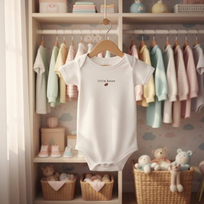 K&ouml;nnte beinhalten: Ein wei&szlig;er Baby-Body mit dem Text "little bean" und einer kleinen braunen Bohne. Der Body h&auml;ngt an einem Holzb&uuml;gel in einem Kinderzimmerschrank voller Babykleidung und Spielzeug.