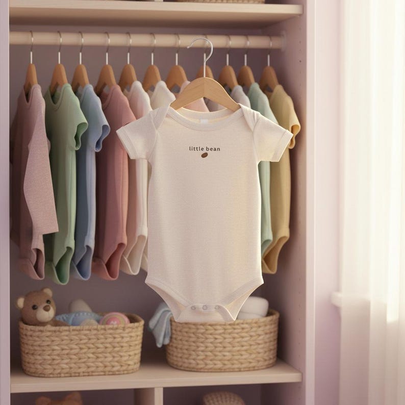 K&ouml;nnte beinhalten: Ein wei&szlig;er Baby-Body mit dem Text "little bean" und einer kleinen braunen Bohne. Der Body h&auml;ngt an einem Holzb&uuml;gel in einem Schrank voller Babykleidung in Pastellfarben. Darunter befinden sich Weidenk&ouml;rbe.