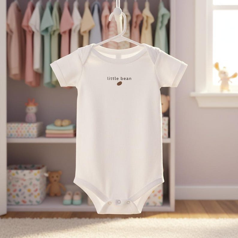 K&ouml;nnte beinhalten: Wei&szlig;er Baby-Body mit kurzen &Auml;rmeln, h&auml;ngt an einem wei&szlig;en Kleiderb&uuml;gel. Auf dem Body steht in brauner Schrift "little bean". Im Hintergrund ist ein Schrank mit Babykleidung und Spielzeug zu sehen.