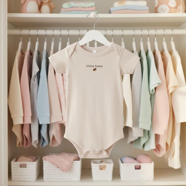 May include: A beige baby onesie with the text "little bean" and a brown bean graphic is hanging on a white hanger. The onesie is surrounded by other baby clothes in pastel colors, including pink, blue, and cream, hanging in a closet.