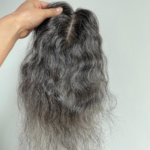 Puede incluir: Una pieza de cabello gris ondulado con una raya de aspecto natural. La pieza capilar se sostiene para mostrar la textura y las variaciones de color, con raíces más oscuras y largos grises más claros. El cabello parece suave y voluminoso.