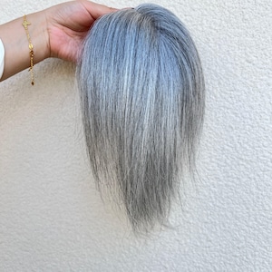 May include: A silver-gray hair topper with long, straight strands. The hairpiece is held up against a white background, showcasing its color and texture. The hairpiece appears to be made of natural hair.
