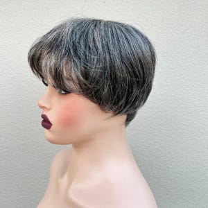 Könnte beinhalten: Ein Schaufensterpuppenkopf mit einer kurzen, gestuften Perücke in Grau- und Schwarztönen. Die Perücke hat einen seitlich gefegten Pony und ist eng am Kopf anliegend gestylt. Die Schaufensterpuppe hat einen hellen Teint mit rosigen Wangen und dunklem Lippenstift.