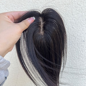 Op de afbeelding: Een donkerbruine haartop met een natuurlijk ogende scheiding. Het haarstuk wordt door een hand omhoog gehouden, waarbij de fijne lokken en het realistische uiterlijk van het haar worden getoond. Het haar lijkt glad en glanzend.