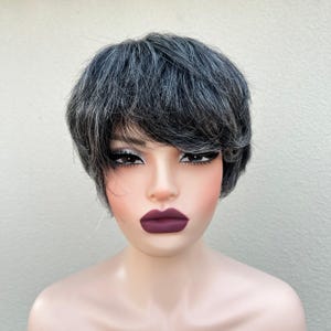 May include: A short, layered wig with a mix of gray and black hair. The wig has a side-swept bang and is styled on a mannequin head. The mannequin has dark lipstick and dramatic eye makeup.