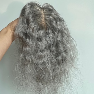 May include: A silver-grey hair topper with wavy, shoulder-length hair. The hairpiece has a natural-looking parting and is designed to add volume and coverage. The hair is a blend of grey and silver tones.