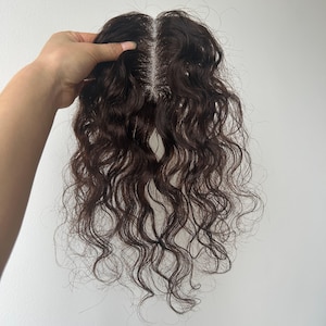 May include: A dark brown hair topper with wavy texture. The hairpiece has a central parting and is held up against a white background. The hairpiece is designed to add volume and coverage to the top of the head.