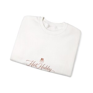 Peut inclure: Un sweat-shirt blanc à col rond avec l'inscription "Hot Hubby" en écriture cursive. Au-dessus du texte, un petit dessin de chapeau haut de forme et de lunettes de couleur cuivre. Le sweat-shirt est plié proprement.