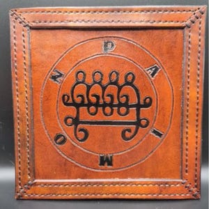 May include: Square, brown leather item with a stitched border. The center features a circular design with an intricate black symbol and letters. The leather has a textured appearance, and the stitching is visible around the edges.