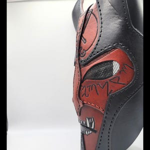 Op de afbeelding: Close-up van een zwart en rood leren masker met ingewikkelde details. Het masker heeft een gestileerd gezichtsontwerp met een gaas ooggebied en gestikte accenten. Het kleurenschema is voornamelijk zwart en rood, met witte accenten.