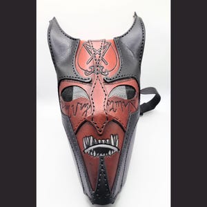 May include: A handcrafted leather mask in a devil design. The mask features a red face with black horns and accents, mesh eye coverings, and a strap. The mask has a stitched design and a detailed mouth with white teeth.