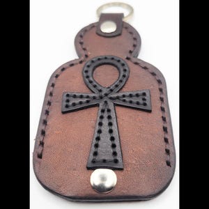 May include: Brown leather keychain featuring a black Ankh symbol. The keychain has a metal ring and a snap closure at the bottom. The Ankh symbol is stitched onto the leather.