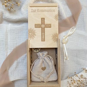 Puede incluir: Una caja de madera con tapa con bisagras, grabada con una cruz y las palabras "Zur Kommunion". En el interior, una pequeña bolsa de arpillera con un diseño de corazón descansa sobre un lecho de virutas de madera. Una cinta color crema está atada a la caja.