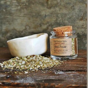 May include: A small glass jar with a cork lid filled with a blend of herbs and salt. The jar has a label that reads "Alta Cucina Italia", "Flavoured Salt", "Positano", and "www.altacucinitalia.com".