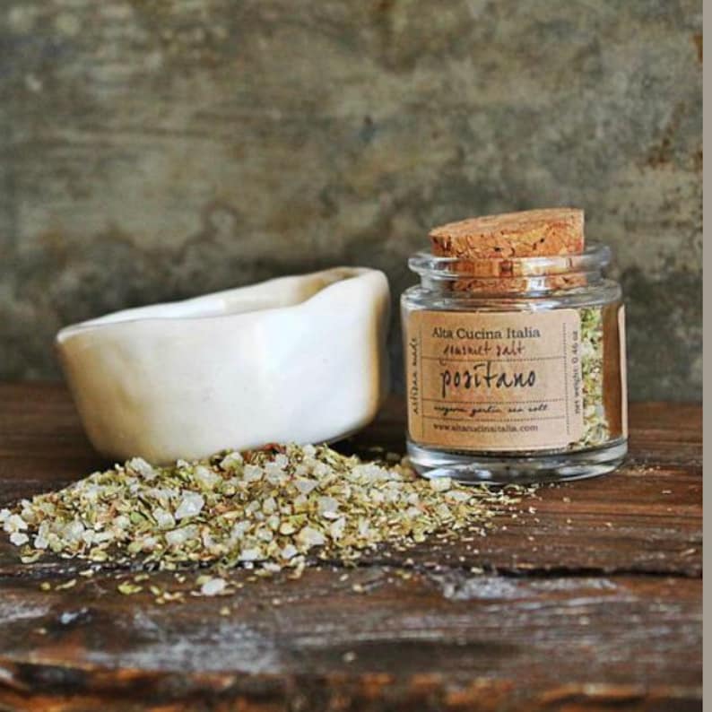 May include: A small glass jar with a cork lid filled with a blend of herbs and salt. The jar has a label that reads "Alta Cucina Italia", "Flavoured Salt", "Positano", and "www.altacucinitalia.com".