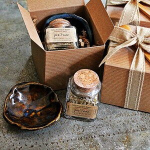 May include: Two small glass jars with cork lids filled with dried herbs and labelled "Alta Cucina Italia -  Positano" are displayed in brown cardboard boxes with a brown ribbon. A small brown ceramic dish is in the foreground.