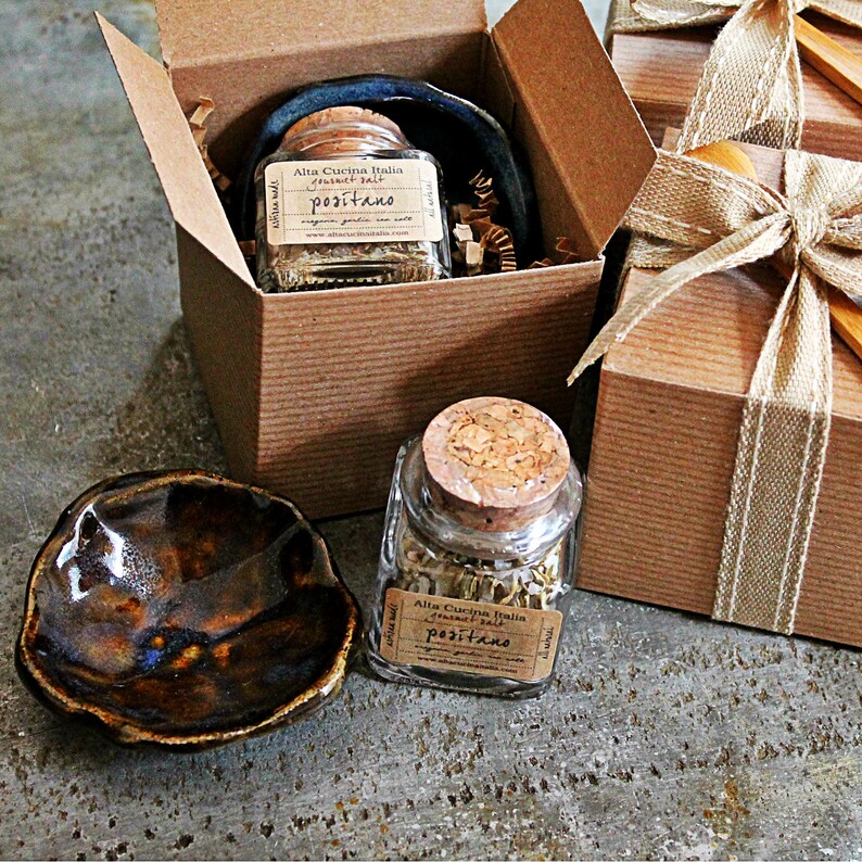 May include: Two small glass jars with cork lids filled with dried herbs and labelled "Alta Cucina Italia -  Positano" are displayed in brown cardboard boxes with a brown ribbon. A small brown ceramic dish is in the foreground.