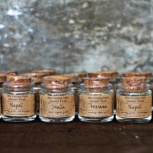 May include: A collection of ten small glass jars with cork lids filled with dried herbs and spices. Each jar has a label with the text "Alta Cucina Italia" and a different region of Italy, including "Halia", "Napoli", "Toscana", and "Romagna".