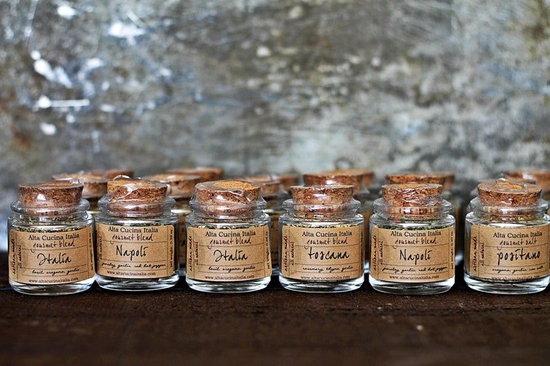 May include: A collection of ten small glass jars with cork lids filled with dried herbs and spices. Each jar has a label with the text "Alta Cucina Italia" and a different region of Italy, including "Halia", "Napoli", "Toscana", and "Romagna".