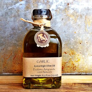 Może przedstawiać: Przezroczysta szklana butelka z oliwą z oliwek extra virgin o smaku czosnkowym z brązową papierową etykietą. Etykieta zawiera napis "GARLIC Extra Virgin Olive Oil Prodotto Artigianale 100% Naturale Net Weight: 8.5 Fl.oz (251ml)"