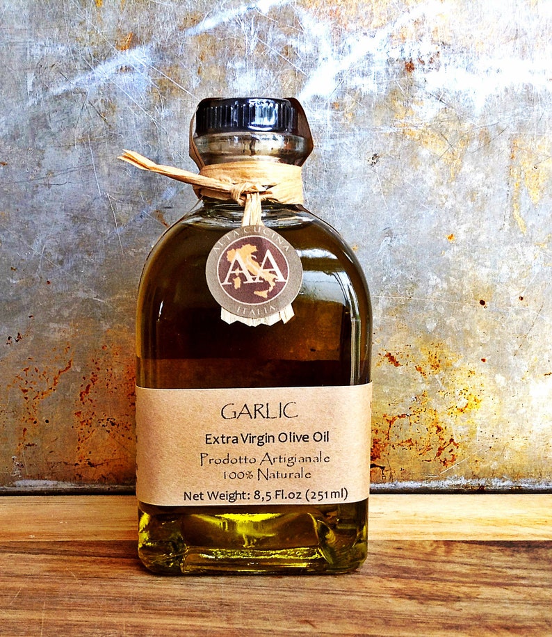 Może przedstawiać: Przezroczysta szklana butelka z oliwą z oliwek extra virgin o smaku czosnkowym z brązową papierową etykietą. Etykieta zawiera napis "GARLIC Extra Virgin Olive Oil Prodotto Artigianale 100% Naturale Net Weight: 8.5 Fl.oz (251ml)"