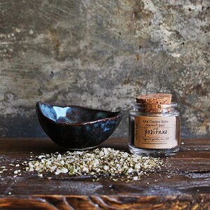 May include: A small glass jar with a cork lid filled with dried herbs and labelled "Ala Cucina Italiana Natural Salt - Positano" sits on a wooden surface next to a small blue and brown ceramic bowl and a scattering of dried herbs.