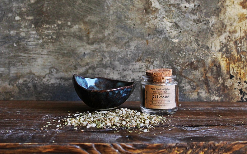 May include: A small glass jar with a cork lid filled with dried herbs and labelled "Ala Cucina Italiana Natural Salt - Positano" sits on a wooden surface next to a small blue and brown ceramic bowl and a scattering of dried herbs.