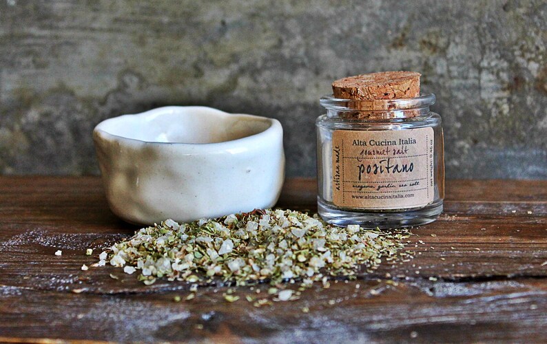 May include: A small glass jar with a cork lid filled with a blend of herbs and salt. The jar has a label that reads "Alta Cucina Italia", "Sea Salt", "Positano", "Oregano Garden Sea Salt", and a website address. The jar is sitting on a wooden surface with a white ceramic bowl and a pile of the herb and salt blend.