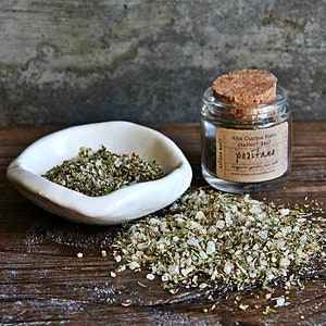 May include: A glass jar with a cork lid filled with a blend of salt and herbs. The label reads "Alta Cucina Italia", "Natural Salt", "Positano", and a website address. The jar is sitting on a wooden surface with a small white dish filled with the same salt and herb blend.