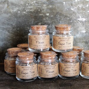 May include: A collection of small glass jars with cork lids filled with gourmet salt blends. The jars are labelled with the names "Firenze" and "Cinque Tese".