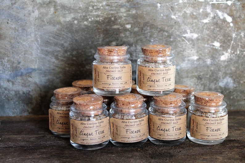 May include: A collection of small glass jars with cork lids filled with gourmet salt blends. The jars are labelled with the names "Firenze" and "Cinque Tese".