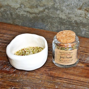 May include: A small glass jar with a cork lid filled with green herbs and a label that reads "Alta Cucina Italia Natural Salt Positano". The jar is sitting on a wooden surface next to a white ceramic bowl filled with a mixture of herbs and salt.