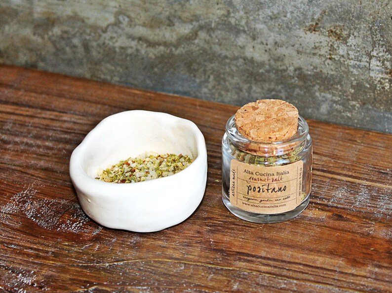 May include: A small glass jar with a cork lid filled with green herbs and a label that reads "Alta Cucina Italia Natural Salt Positano". The jar is sitting on a wooden surface next to a white ceramic bowl filled with a mixture of herbs and salt.