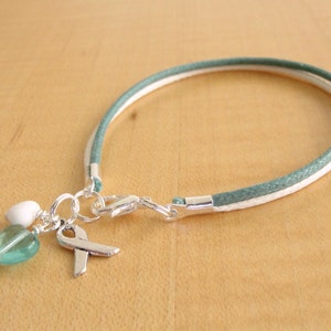 Puede incluir: Una pulsera trenzada de color verde azulado y blanco con un dije de plata con forma de cinta y una cuenta en forma de corazón.
