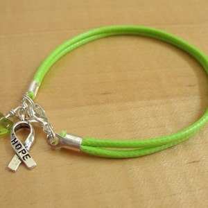 Puede incluir: Un brazalete de cordón verde con un dije de plata con una cinta con la palabra "HOPE" y un dije de corazón verde.