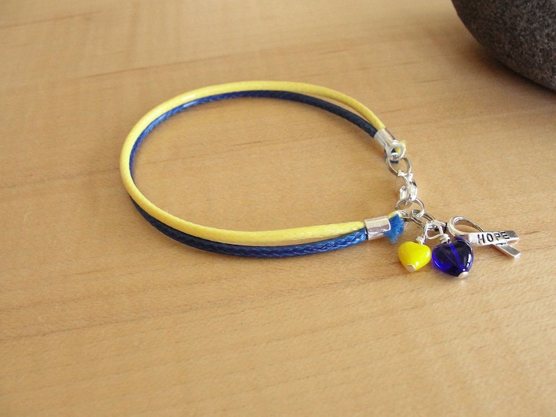 May include: A yellow and blue braided bracelet with a silver charm that says "HOPE" and two heart-shaped beads, one yellow and one blue.