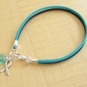 May include: Teal and black cord bracelet with a silver charm featuring a teal heart and a silver ribbon.