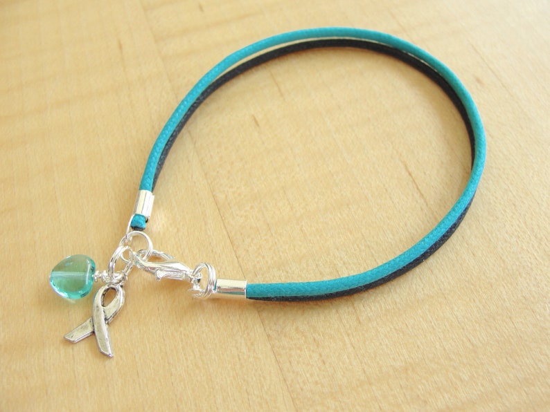 May include: Teal and black cord bracelet with a silver charm featuring a teal heart and a silver ribbon.