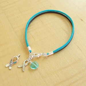 May include: Teal and black cord bracelet with a silver charm featuring a ribbon with the word "HOPE" and a teal glass bead.
