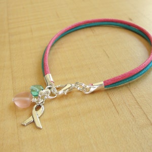 May include: A pink and teal cord bracelet with a silver charm featuring a pink and green glass bead and a silver ribbon.