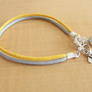 May include: A yellow and gray braided bracelet with a silver charm featuring a ribbon with the word "HOPE" and a silver bead.