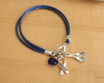 Puede incluir: Una pulsera de doble cordón azul oscuro con tres dijes de metal plateado: una cuenta de vidrio azul en forma de corazón, un dije en forma de cinta con la palabra HOPE grabada y dos pequeños dijes en forma de cuchara unidos a un broche.