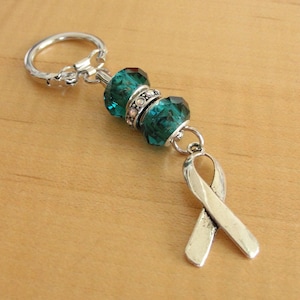 Peut inclure: Un porte-clés argenté avec un accent de perles de couleur teal et un charm en forme de ruban. Le porte-clés comporte un anneau décoratif et un fermoir mousqueton. Les perles teal sont facettées et ont un aspect cristallin.